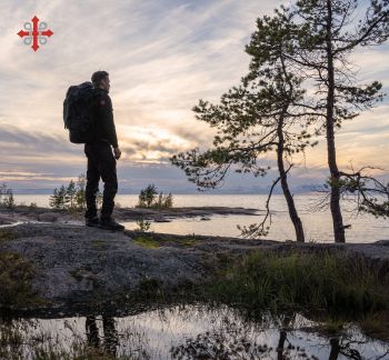 Vandrare med ryggsäck står på en klippa vid havet i solnedgång, med tallar i förgrunden och St. Olav‑symbolen i hörnet.